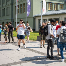 A group of students is gathered outside a building labeled "Hochschule Anhalt University of Applied Sciences." Some students are walking, while others are standing and talking. The scene appears to be on a sunny day, with people dressed in casual attire.