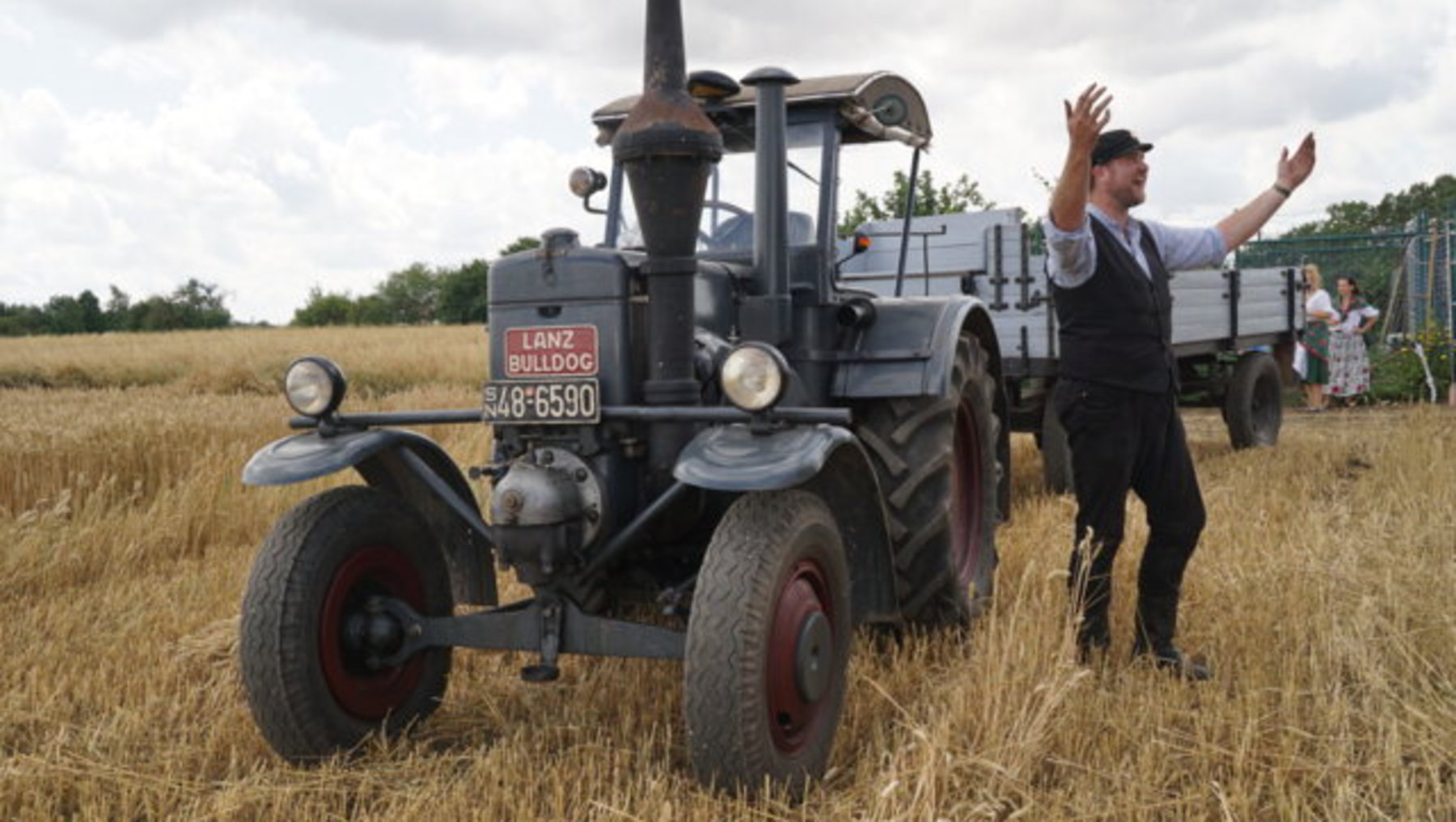 Ein alter Lanz Bulldog-Traktor mit rotem Kennzeichen steht auf einem Feld mit hohem Stroh. Ein Mann in historischer Kleidung winkt neben dem Fahrzeug, während im Hintergrund weitere Personen zu sehen sind.