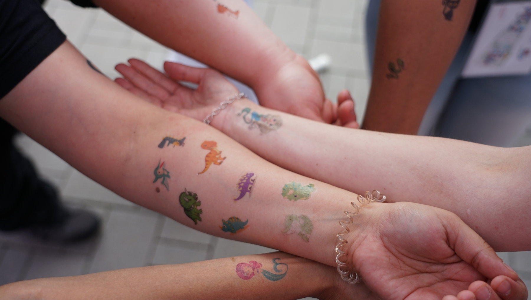 Mehrere Arme mit bunten, kleinen Dinosaurier-Tattoos in verschiedenen Farben und Stilen, die sich gegenseitig berühren oder stützen.