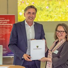 Ein Mann und eine Frau stehen lächelnd nebeneinander und überreichen sich einen Ordner mit dem Logo „Sächsische Energie“. Die Frau hält zusätzlich eine Urkunde.