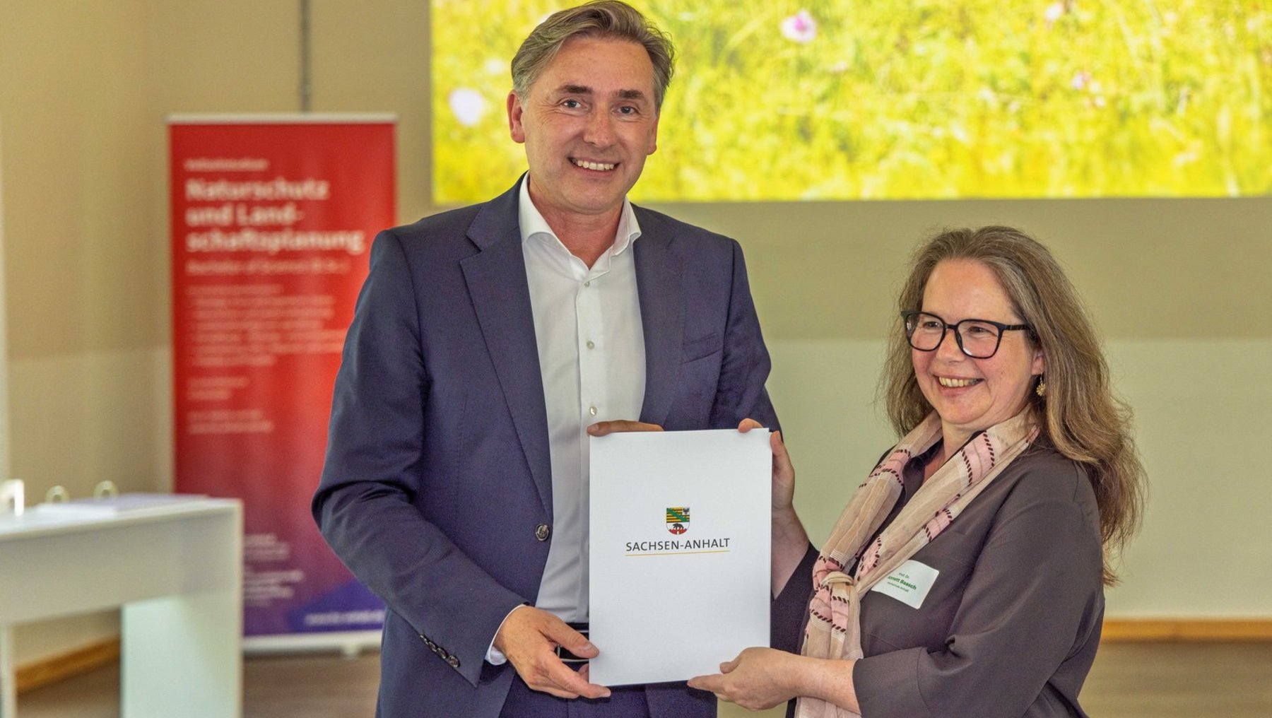 Ein Mann und eine Frau stehen lächelnd nebeneinander und überreichen sich einen Ordner mit dem Logo „Sächsische Energie“. Die Frau hält zusätzlich eine Urkunde.
