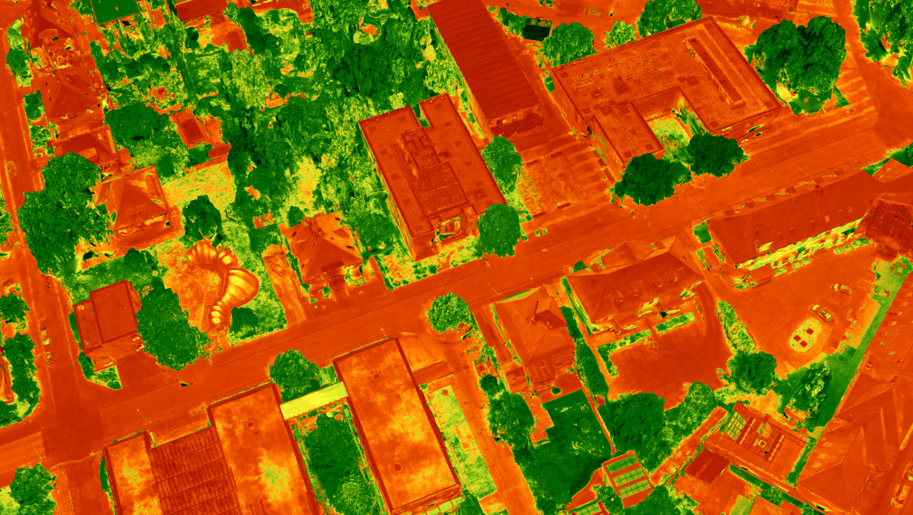 Luftaufnahme eines Stadtgebiets in Wärmebilddarstellung mit Gebäuden, Straßen und Grünflächen, die sich durch unterschiedliche Farbintensitäten (rot/orange für wärmere Bereiche, grün für kühlere) abzeichnen.