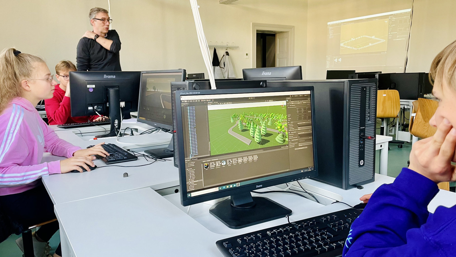 Drei Kinder arbeiten in einem Computerraum an Bildschirmen mit einer 3D-Software, die eine grüne Landschaft oder ein Grundrissmodell zeigt. Eine Lehrperson beobachtet im Hintergrund.