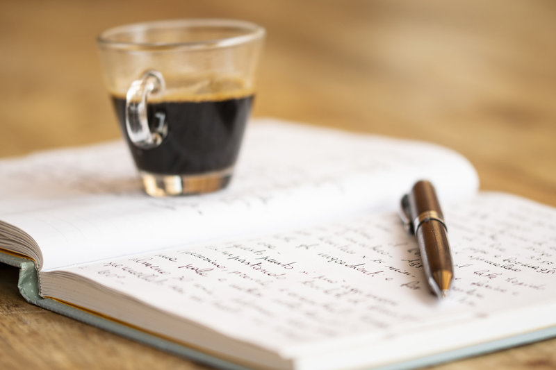 Auf einem Tisch steht eine Tasse mit dunklem, heißem Getränk, vermutlich Kaffee. Daneben liegt ein offenes Notizbuch mit handschriftlichen Notizen und ein Füllfederhalter. Ein Buch ist teilweise sichtbar.