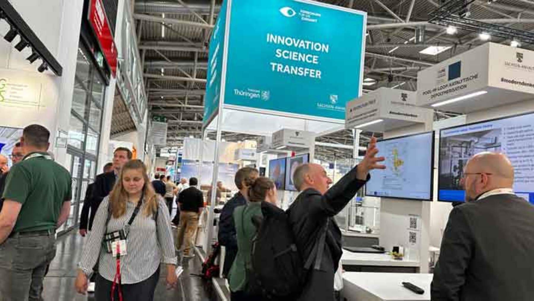 Messebesucher auf einer Ausstellung zu „Innovation, Science, Transfer“: Ein Mann zeigt auf eine große blaue Infotafel, während andere Besucher in der Halle um ihn herumstehen und sich unterhalten.