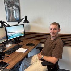 Ein lächelnder Mann sitzt an einem Schreibtisch mit zwei Bildschirmen, die eine Küstenlandschaft zeigen. Vor ihm liegen Tastatur, Maus und ein Notizblock. Auf dem Tisch stehen eine Tasse und ein Glas.