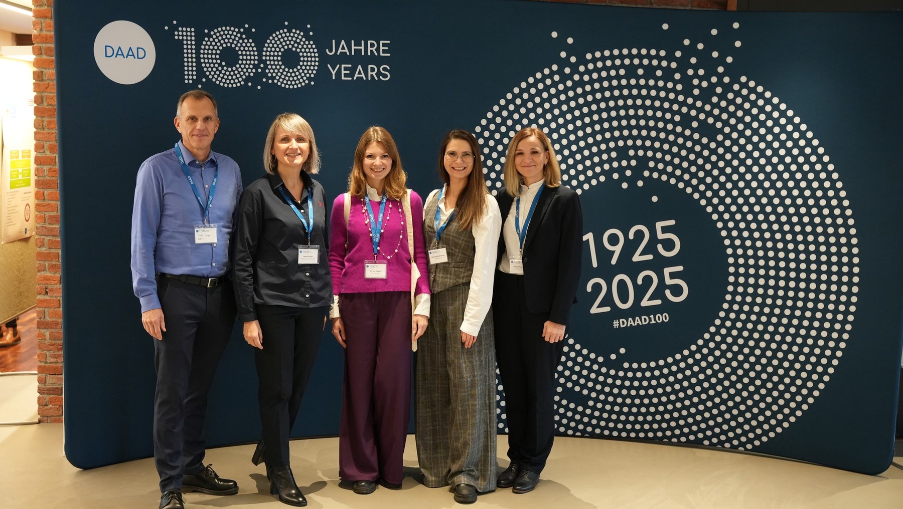 "Fünf Personen stehen vor einem blauen Hintergrund, der das 100-jährige Jubiläum des DAAD (Deutscher Akademischer Austauschdienst) von 1925 bis 2025 feiert. Die Personen lächeln und tragen Namensschilder. Der Hintergrund zeigt das DAAD-Logo und den Hashtag #DAAD100."