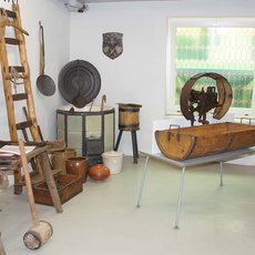 Ein Raum mit alten Werkzeugen und Geräten, darunter eine Leiter, ein Webstuhl, verschiedene Holzgegenstände, Metallschilder und andere traditionelle Handwerksutensilien. Die Gegenstände sind auf Tischen und Regalen angeordnet, mit einem Fenster im Hintergrund, das auf einen Garten hinausgeht.