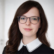 Porträt einer Frau mit mittellangen, braunen Haaren, die eine runde Brille trägt. Sie lächelt leicht und trägt ein schwarzes Oberteil mit weißem Stehkragen.