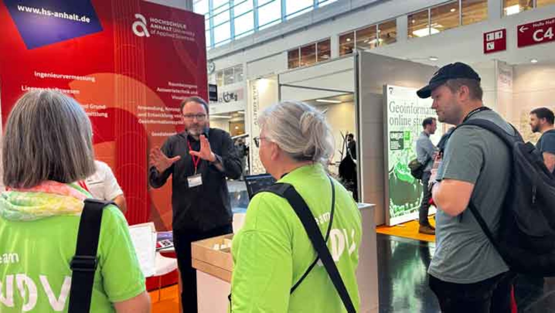 Drei Personen auf einer Messe stehen vor einem Infostand der Hochschule für angewandte Wissenschaften. Ein Mann in Business-Kleidung erklärt mit Gesten etwas, während zwei Frauen in neonfarbenen Westen mit Rucksäcken zuhören. Im Hintergrund sind weitere Aussteller und Infomaterialien zu sehen.