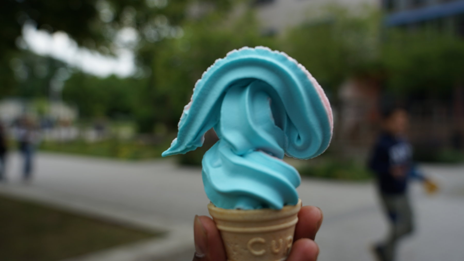 Ein Hand hält ein Eis in Form eines blauen Delfins mit weißer Unterseite in einem Waffelbecher mit der Aufschrift „CU“.