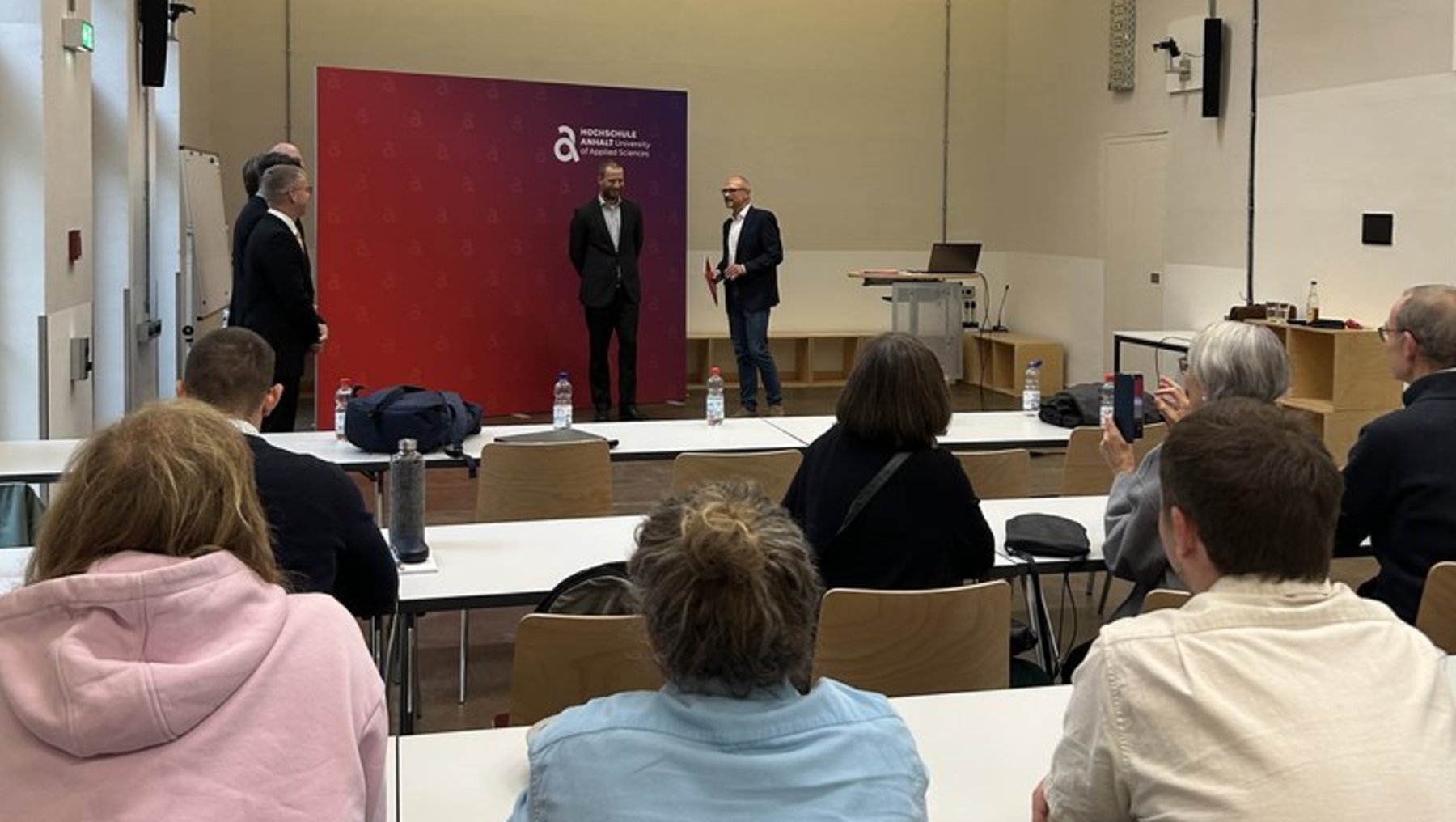 Vier Personen stehen auf einer Bühne vor einem roten Hintergrund und halten eine kurze Ansprache. Im Saal sitzen etwa 15 Zuhörer an Tischen, die ihnen zugewandt sind. Die Atmosphäre wirkt formell, etwa bei einer Veranstaltung oder Präsentation.