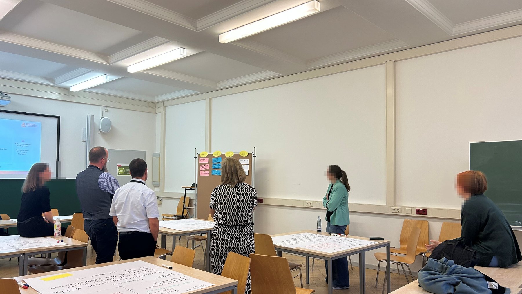 Gruppe von sechs Personen in einem hellen Klassenzimmer mit Holzschultischen und Stühlen. Einige stehen im Kreis, andere sitzen. An der Wand ein Whiteboard mit Notizen und ein Beamer mit Präsentation. Eine Person hält ein Plakat mit farbigen Karten.
