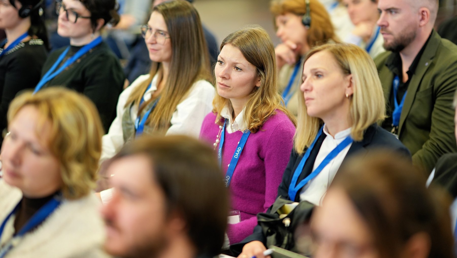 Eine Gruppe von Menschen sitzt in einem Raum und hört aufmerksam einer Präsentation oder einem Vortrag zu. Die meisten tragen Namensschilder und wirken konzentriert. Es scheint sich um eine Konferenz oder ein Seminar zu handeln.