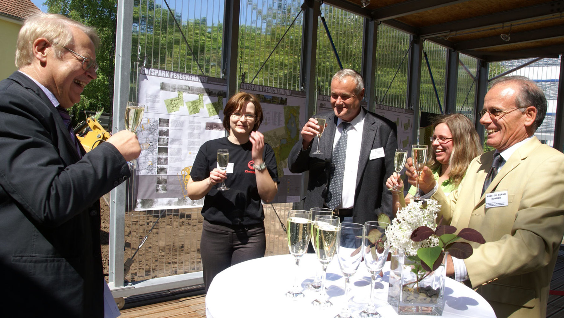 Eine Gruppe von fünf Personen steht um einen Tisch mit Gläsern und Blumen. Sie halten Sektgläser in den Händen und scheinen anzustoßen. Im Hintergrund ist eine Glaswand mit einer Pflanzenskizze zu sehen. Die Atmosphäre wirkt feierlich und freundlich.