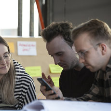 Drei Personen sitzen an einem Tisch, konzentriert auf ein Smartphone schauend, das eine Frau hält, während zwei Männer daneben auf den Bildschirm blicken.