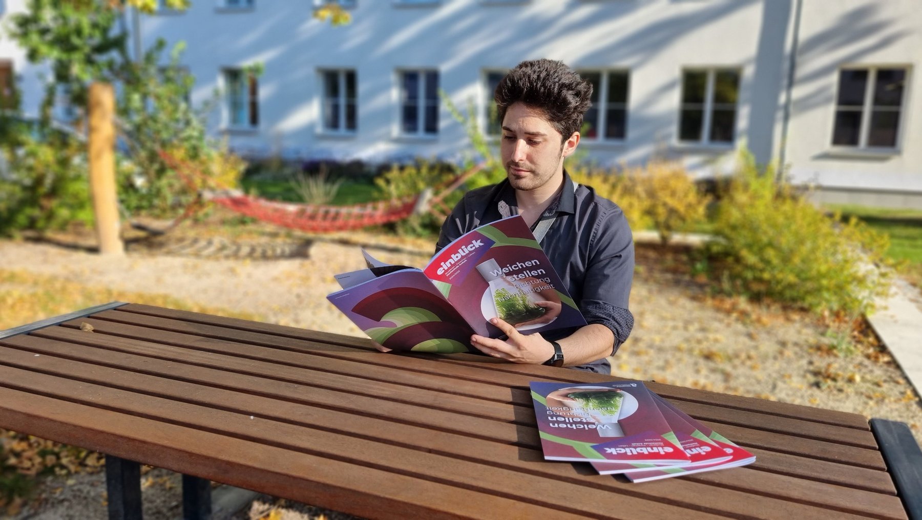 Ein Mann sitzt auf einer Bank im Freien und liest ein Magazin mit dem Titel "Landwirtschaft". Vor ihm auf dem Tisch liegen weitere Magazine. Im Hintergrund sind Gebäude und Pflanzen zu sehen.