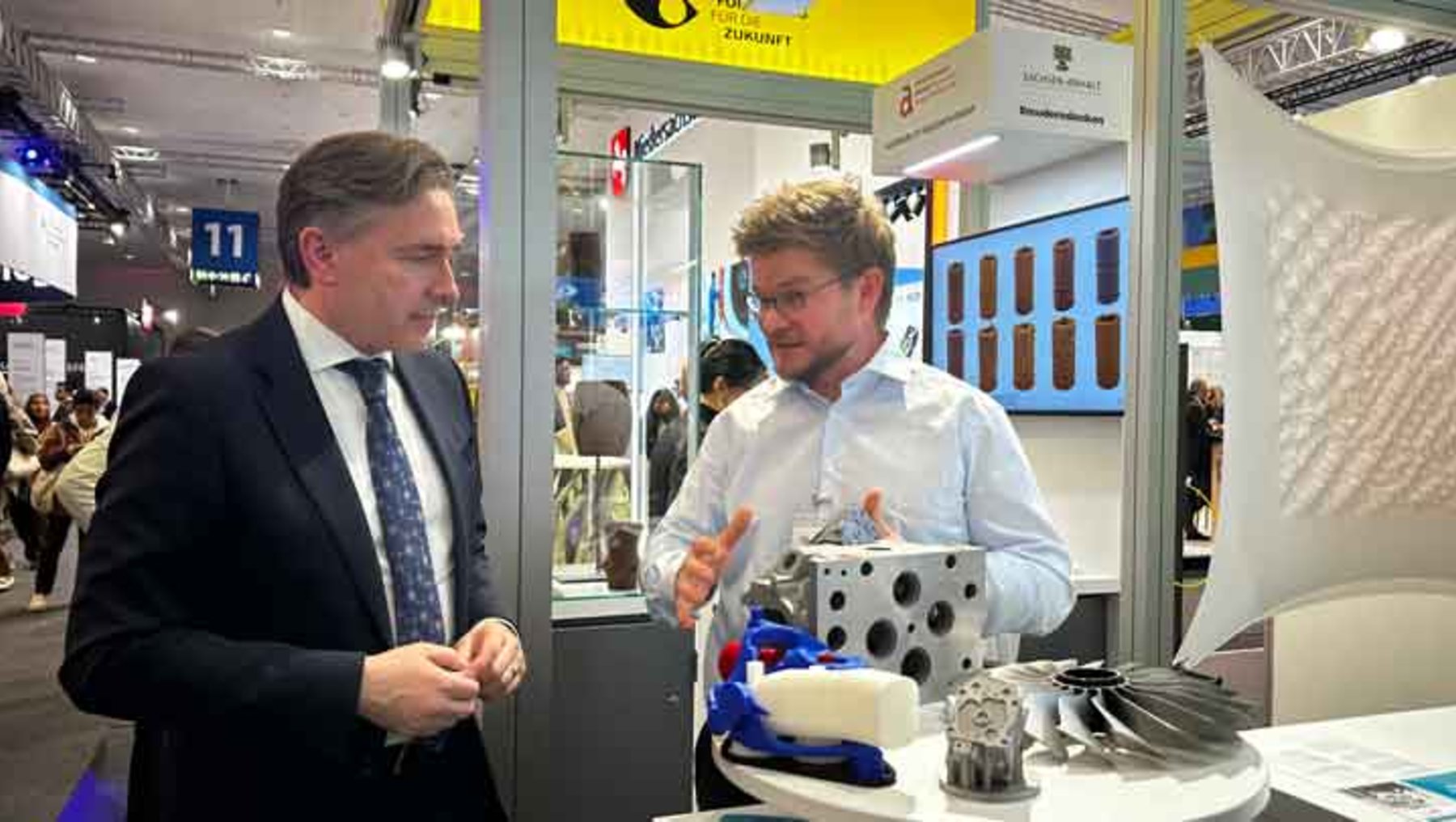 Zwei Männer auf einer Messe: Einer in Anzug, der anderen in Hemd, besprechen ein ausgestelltes technisches Bauteil aus Metall mit strukturierter Oberfläche, umgeben von weiteren Modellteilen.