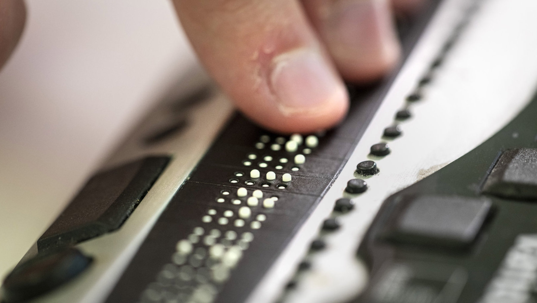 Eine Hand berührt eine Braillezeile, auf der durch Punkte gebildete Buchstaben und Symbole zu erkennen sind.