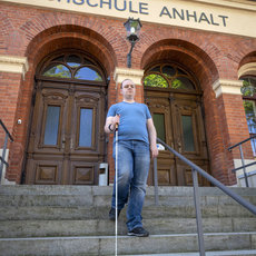 Ein Mann mit weißem Langstock steigt die Stufen vor dem Haupteingang der Hochschule Anhalt hinauf, die durch eine große, braune Tür und den Schriftzug „Hochschule Anhalt“ an der Fassade gekennzeichnet ist.