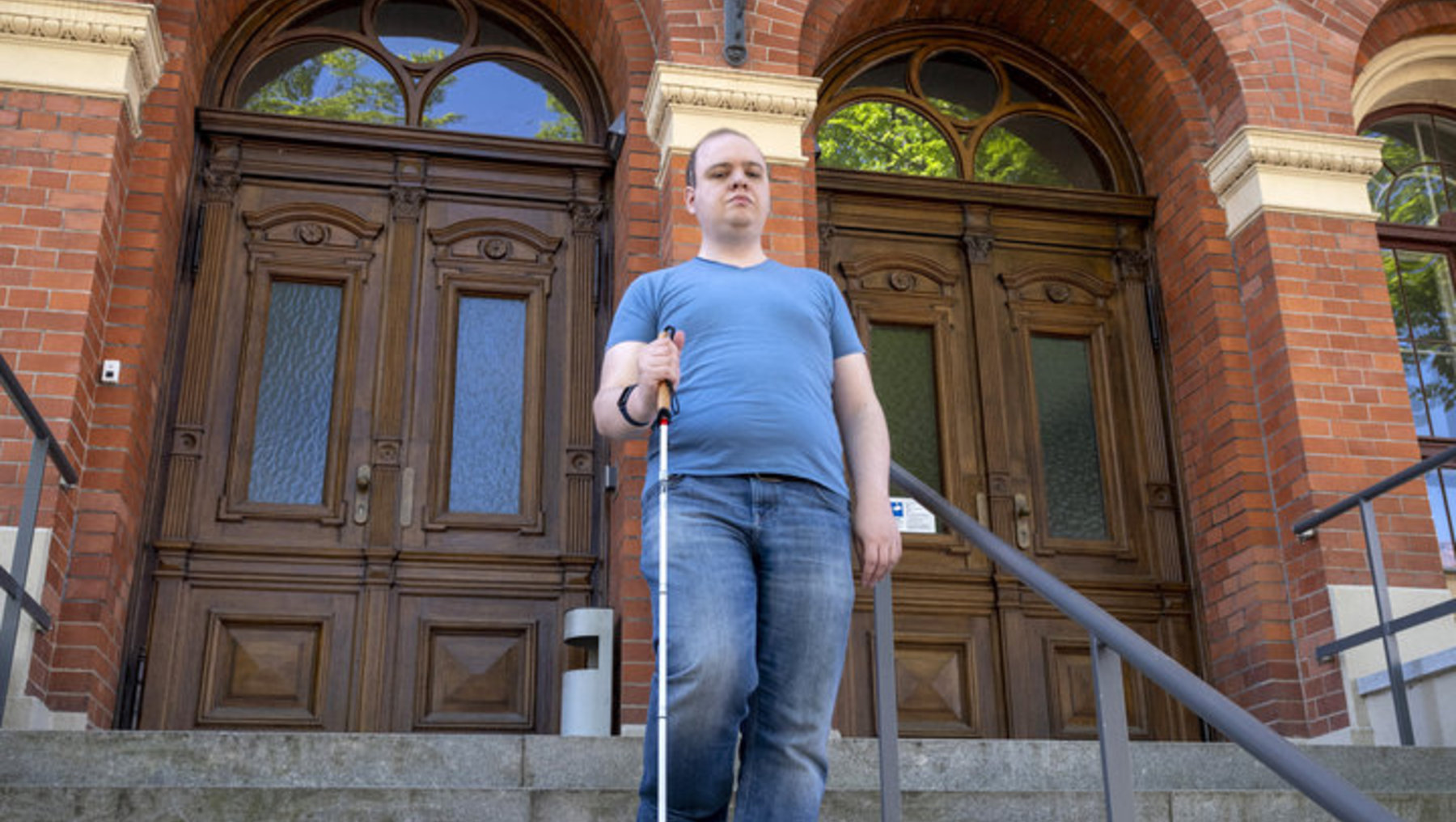 Ein Mann mit weißem Langstock steigt die Stufen vor dem Haupteingang der Hochschule Anhalt hinauf, die durch eine große, braune Tür und den Schriftzug „Hochschule Anhalt“ an der Fassade gekennzeichnet ist.