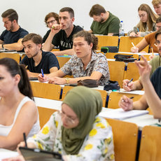 Ein Hörsaal mit Studieninteressierten, die aufmerksam zuhören und Notizen machen. Einige heben die Hand, um Fragen zu stellen. Die Studierenden sind in einer typischen Vorlesungssituation und wirken konzentriert und engagiert.