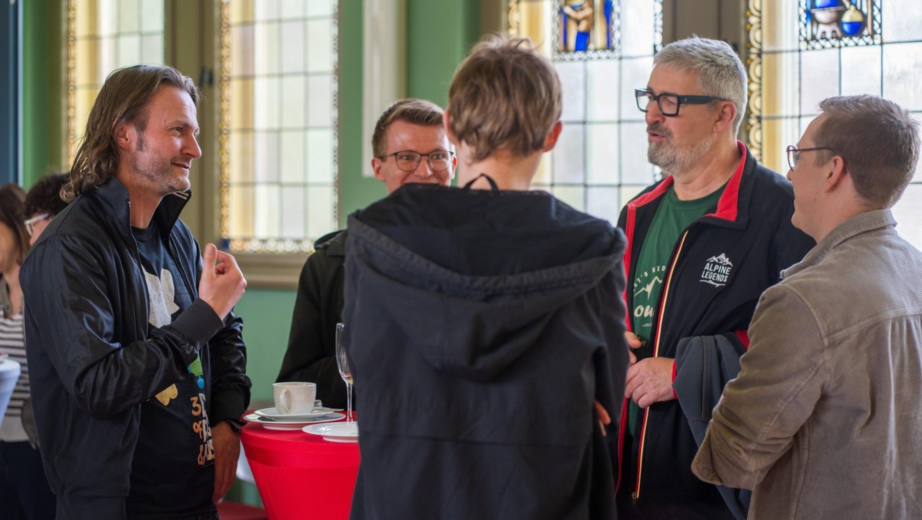 Fünf Männer stehen in einem hellen Raum mit bunten Glasfenstern und unterhalten sich lebhaft im Kreis. Einer hält ein Getränk, ein anderer trägt ein T-Shirt mit Vereinslogo. Im Hintergrund ein Tisch mit Kaffeetasse.