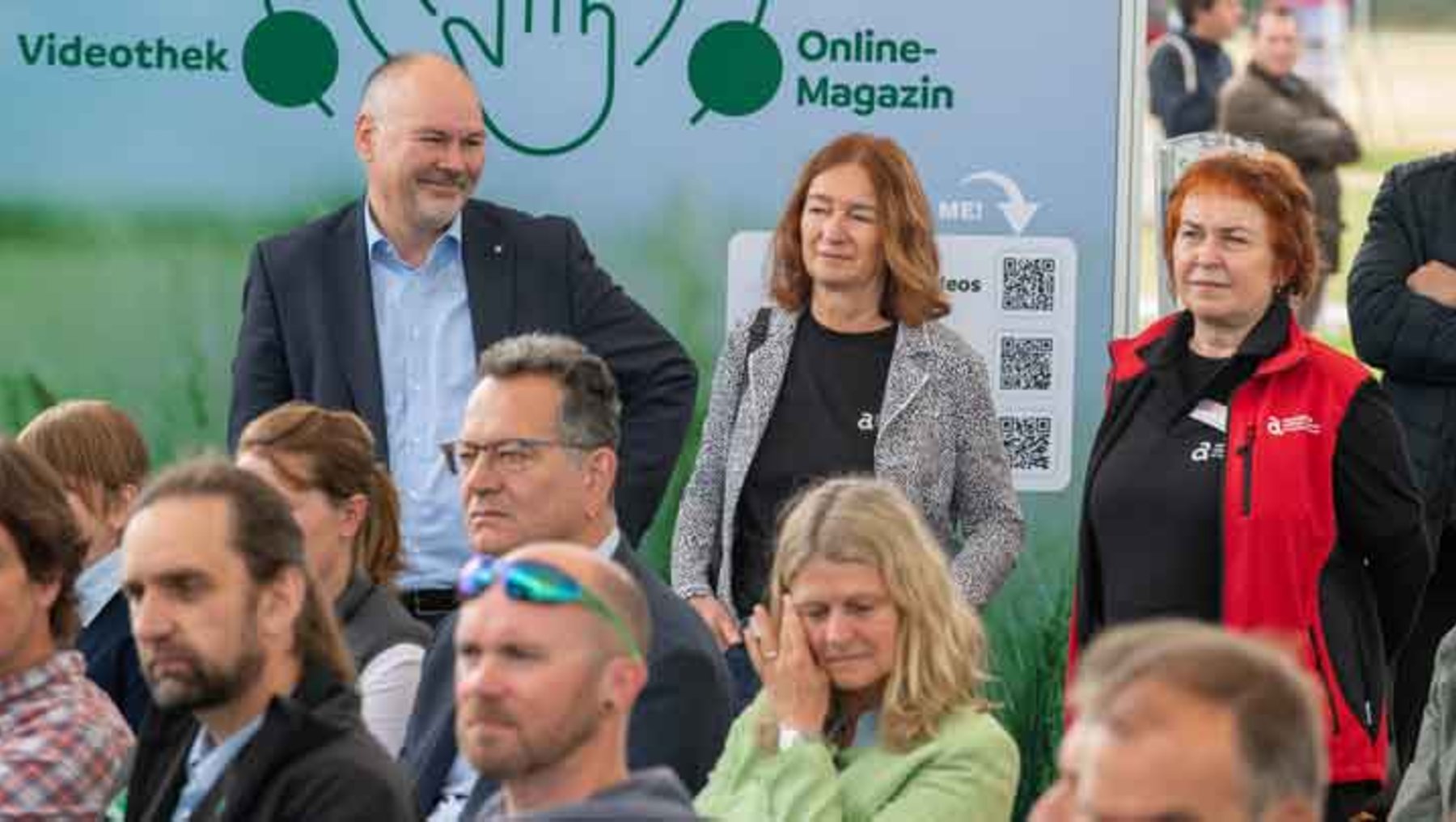 Gruppe von Menschen vor einem Infostand mit dem Schild „Videothek Online-Magazin“, darunter ein QR-Code und ein Logo. Drei Personen im Vordergrund lächeln in die Kamera, während andere im Hintergrund sitzen oder stehen.
