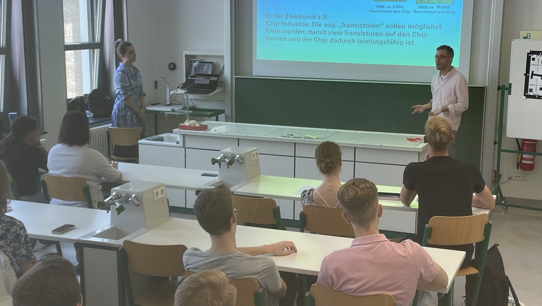 Ein Vortrag im Klassenzimmer: Ein Referent steht vor einer Gruppe von Zuhörern und erklärt am Whiteboard das Thema „Antriebsverfahren“. Auf dem Bildschirm wird die Frage „Welche Antriebsarten gibt es?“ mit Beispielen wie Elektromotoren und Verbrennungsmotoren behandelt.