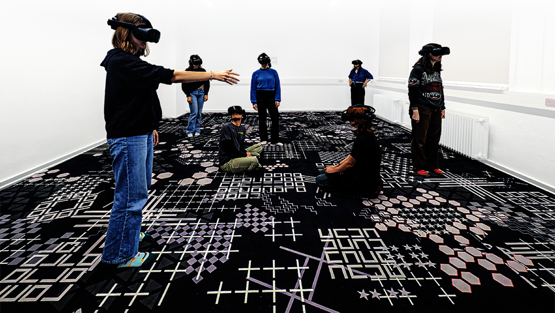 Sieben Personen mit VR-Brillen in einem Raum mit schwarzem, strukturiertem Boden aus geometrischen Mustern und Symbolen. Übungs-VR-Labor für Studierende.