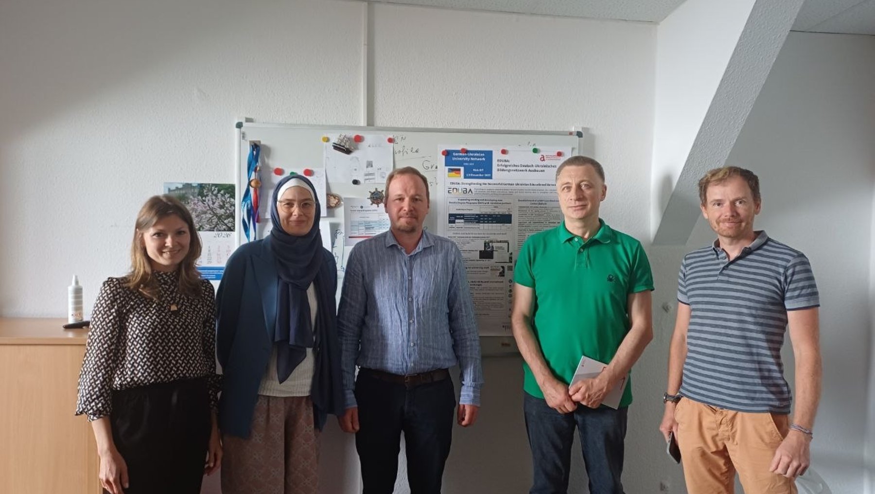 Fünf Personen stehen lächelnd nebeneinander in einem Büro: eine Frau mit langem Haar, eine Frau mit Kopftuch, zwei Männer in Hemden und ein Mann in grünem Shirt mit Krawatte. Im Hintergrund hängt ein Poster mit Diagrammen und Text.