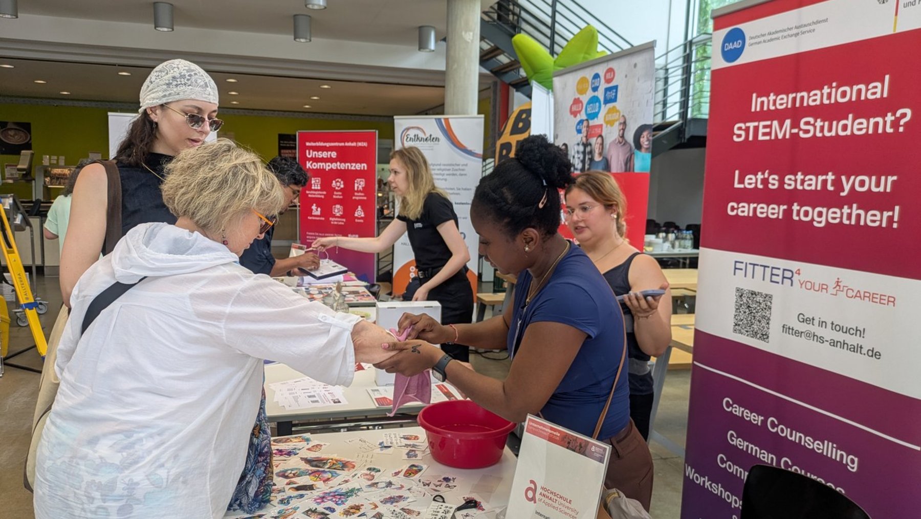 Gruppe von Menschen an einem Infostand zu internationalem STEM-Studium, die gemeinsam ein Poster mit Aufklebern gestalten. Plakat daneben wirbt mit „Internationaler STEM-Student? Lass uns deine Karriere gemeinsam starten!“ und zeigt QR-Code für weitere Infos.