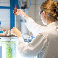 Eine Person in weißem Laborkittel und Schutzbrille hält ein Reagenzglas mit gelber Flüssigkeit und führt ein Experiment in einem Labor durch, während sie eine Apparatur mit grünlicher Flüssigkeit bedient.