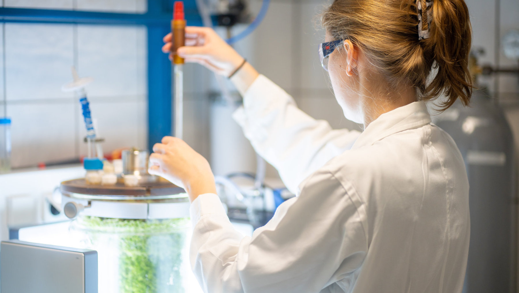 Eine Person in weißem Laborkittel und Schutzbrille hält ein Reagenzglas mit gelber Flüssigkeit und führt ein Experiment in einem Labor durch, während sie eine Apparatur mit grünlicher Flüssigkeit bedient.