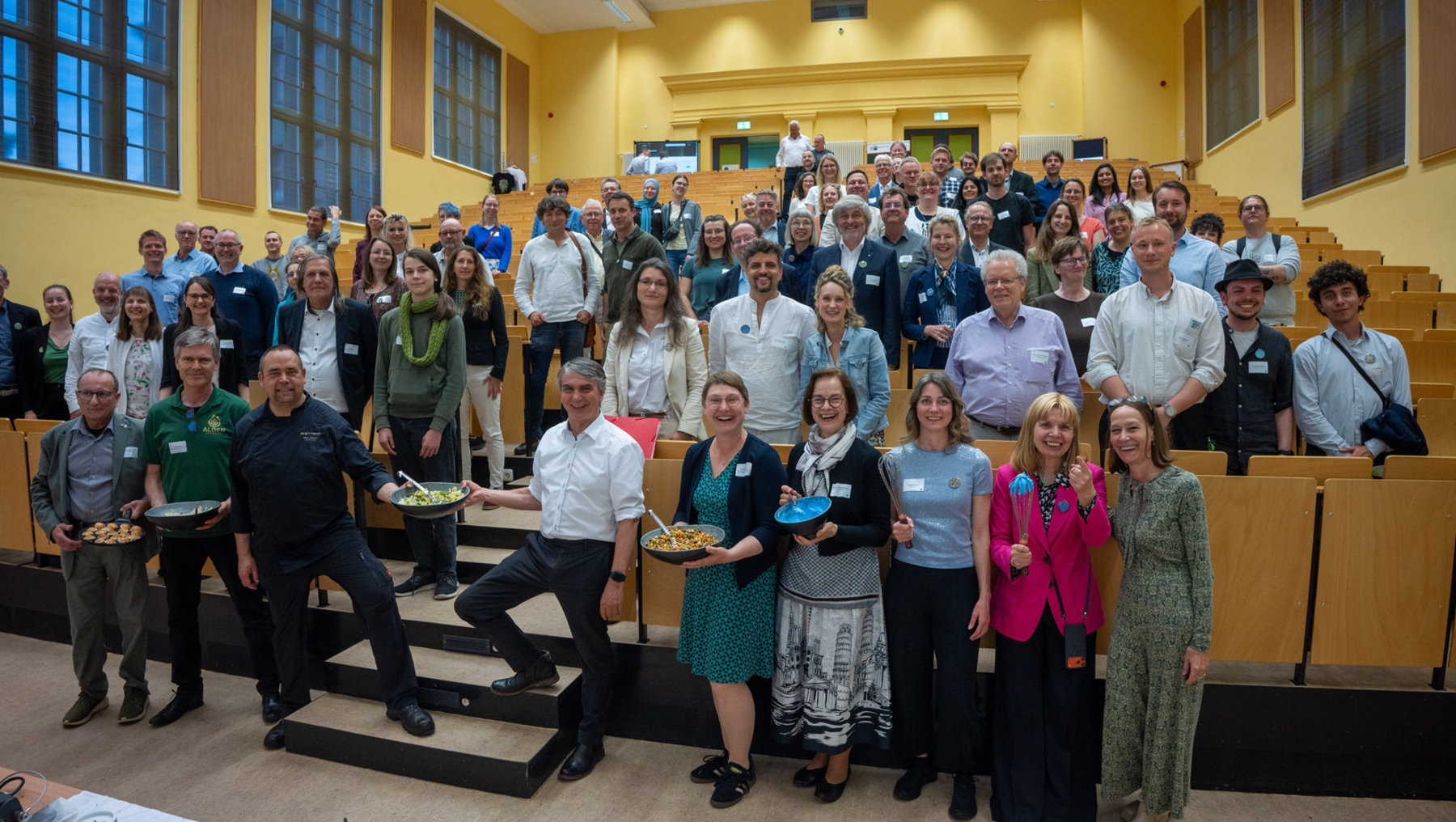 Große Gruppe von etwa 50 Personen, die in einem Hörsaal mit Holzstufen und gelben Wänden versammelt ist. Einige halten Teller mit Snacks, alle wirken gesellig und lächeln für ein Gruppenfoto.