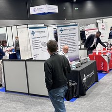 Messe mit mehreren Ausstellungsständen, an denen Besucher mit Ausstellern sprechen. Ein Mann steht vor einem Infotisch mit Plakaten und Flyern zu Themen wie Patientensicherheit und Medizintechnik. Im Hintergrund weitere Stände mit Fachpersonal und Interessenten.