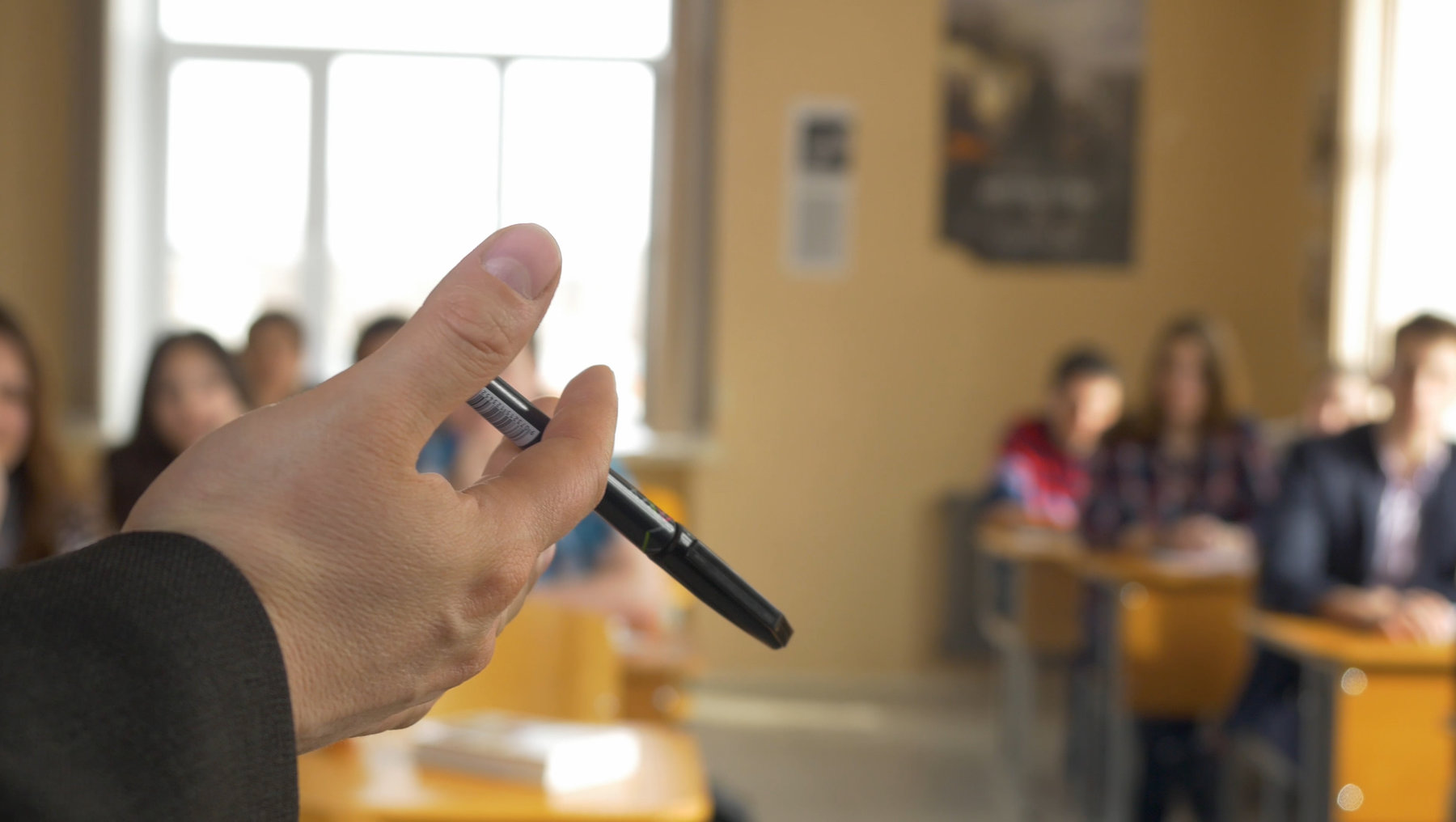 Eine Person hält einen Stift in der Hand und deutet damit auf ein unscharfes Publikum im Hintergrund, das in einem hellen Raum mit Tischen und Stühlen sitzt.