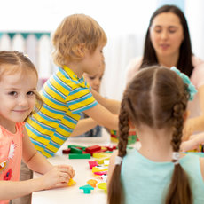 A group of young children are seated at a table, engaged in a craft activity. They are smiling and appear to be enjoying themselves. An adult is supervising them, ensuring they have a fun and safe experience. The room is bright and colorful, creating a cheerful atmosphere.