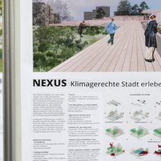 Diese Abbildung zeigt eine Frau, die bei einer Präsentation oder Besichtigung eines nachhaltigen Stadtentwicklungsprojekts, „NEXUS Klimagerechte Stadt erleben“, steht und auf Konzeptzeichnungen eines grünen urbanen Projekts blickt. Es werden modellhafte Beispiele wie Parkanlagen und Wohnräume dargestellt.oxygen_