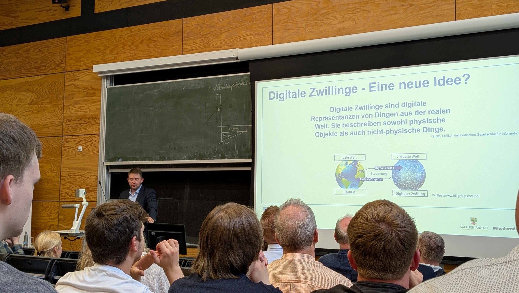 Ein Vortragender steht vor einem Publikum und präsentiert eine Folie zum Thema digitale Zwillinge. Die Folie zeigt den Titel „Digitale Zwillinge – Eine neue Idee?“ und erklärt, dass digitale Zwillinge digitale Repräsentationen realer Objekte sind, die sowohl physische als auch nicht-physische Eigenschaften abbilden.
