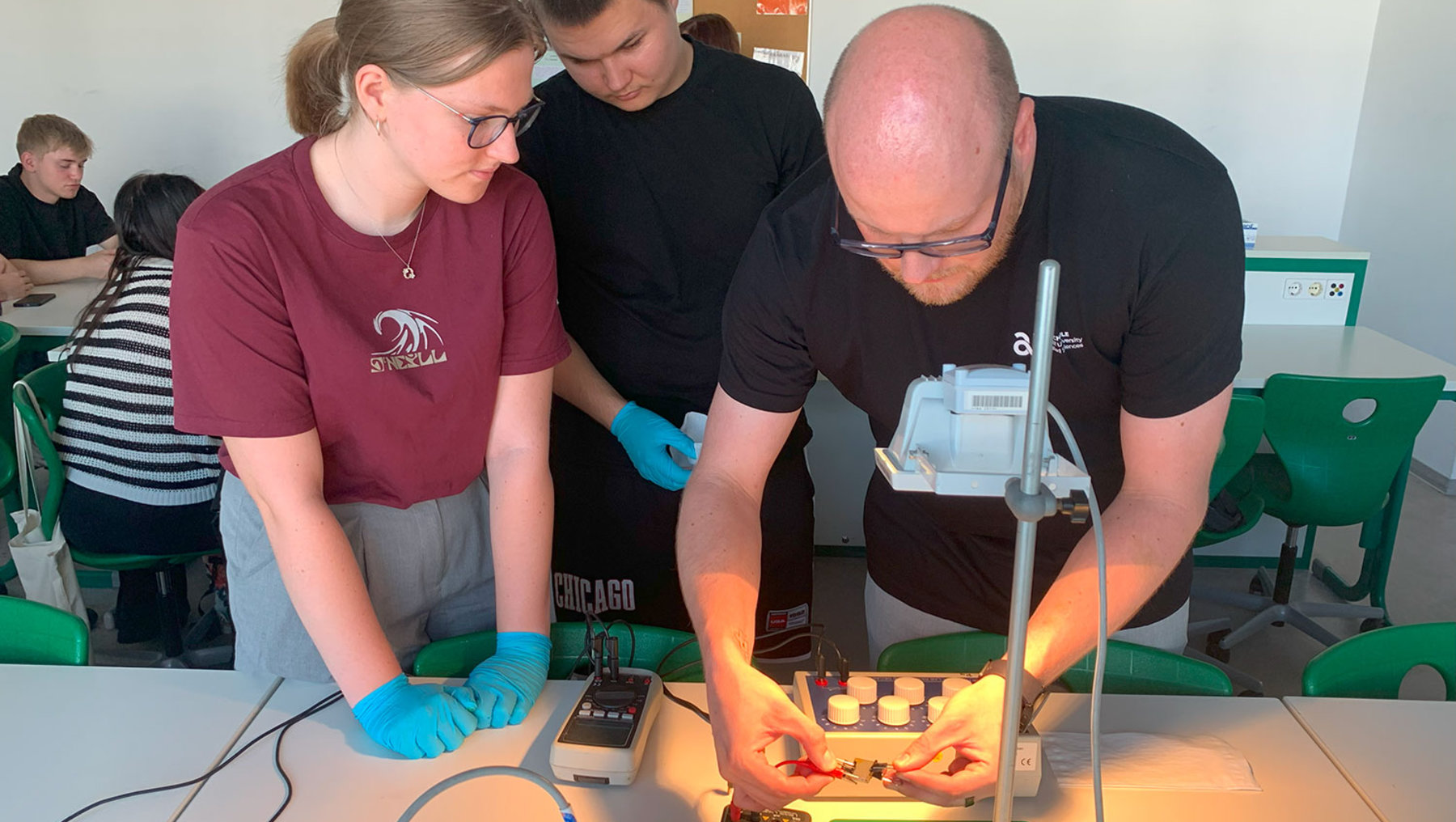 Zwei Personen in Schutzhandschuhen beobachten und arbeiten gemeinsam mit einem Hochschulmitarbeiter an einem Experimentiertisch mit elektronischen Bauteilen und einer leuchtenden, grünen Flüssigkeit in einer Wanne, während sie sich über eine Schaltung beugen.