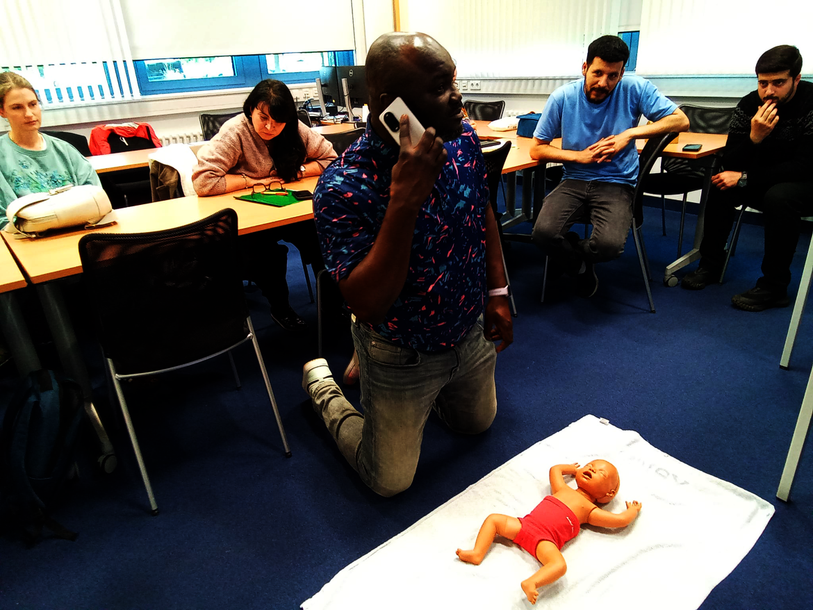 Ein Mann kniet vor einer Gruppe von Menschen in einem Raum und telefoniert, während er eine Puppe in Babykleidung auf einem Tuch hält. Die Gruppe beobachtet die Situation.