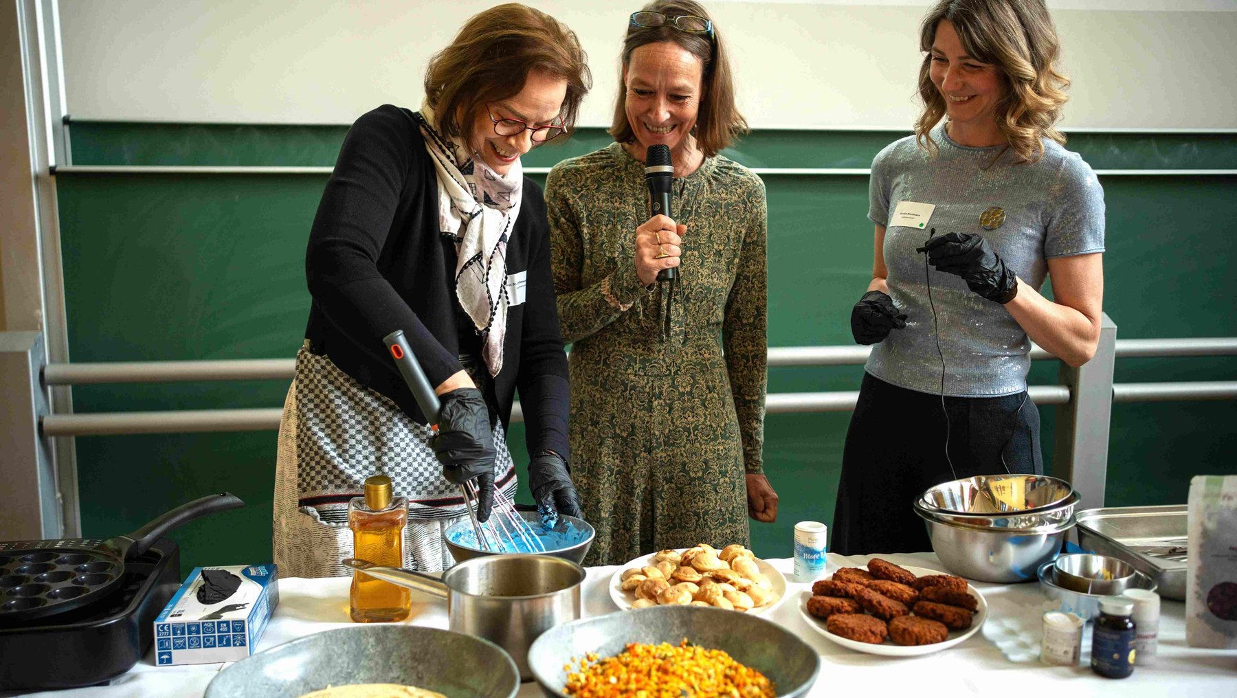 Drei Frauen stehen in einem Hörsaal der Hochschule Anhalt und bereiten gemeinsam ein Buffet vor: eine hält ein Mikrofon und spricht, eine rührt in einem Topf, die dritte hält ein Tablett mit kleinen Snacks. Auf dem Tisch liegen verschiedene Speisen wie Nudeln, Käse und Gebäck.
