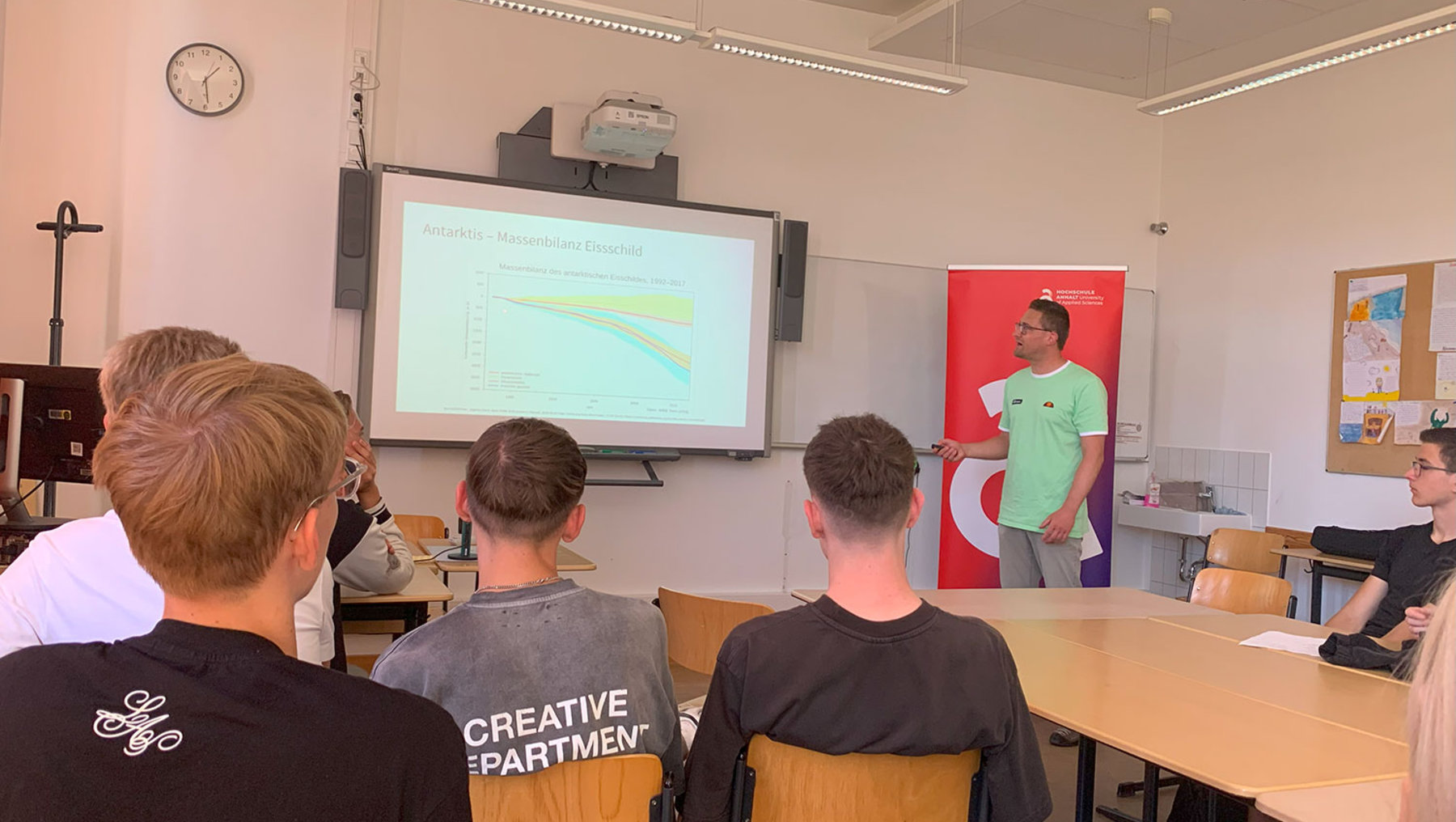 Ein Referent steht vor einem Publikum im Klassenzimmer und präsentiert eine Folie mit einem Diagramm, das einen abfallenden Trend zeigt. Die Zuhörer sitzen an Tischen und folgen der Präsentation.