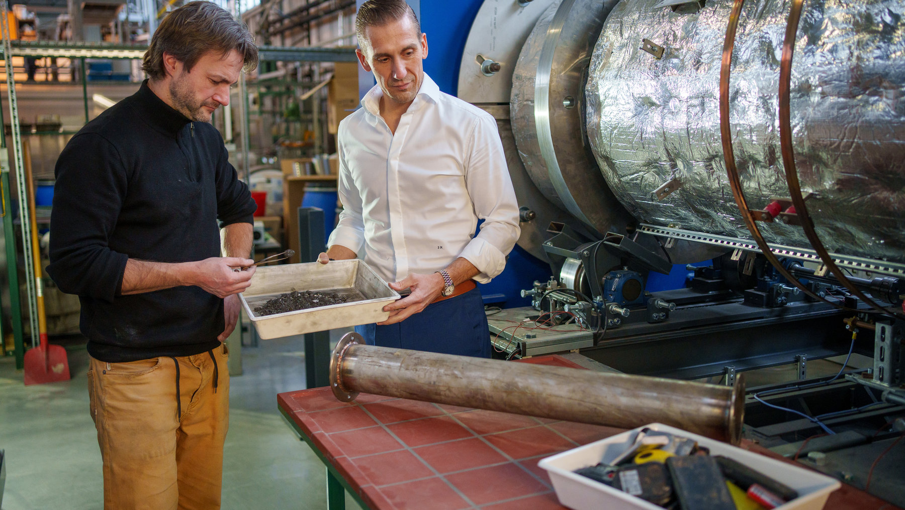 Zwei Männer in einer Werkstatt untersuchen gemeinsam eine offene Kiste mit Werkzeugen. Im Hintergrund steht eine große Maschine mit reflektierender Oberfläche. Sie scheinen sich auf eine technische Aufgabe oder Reparatur zu konzentrieren.