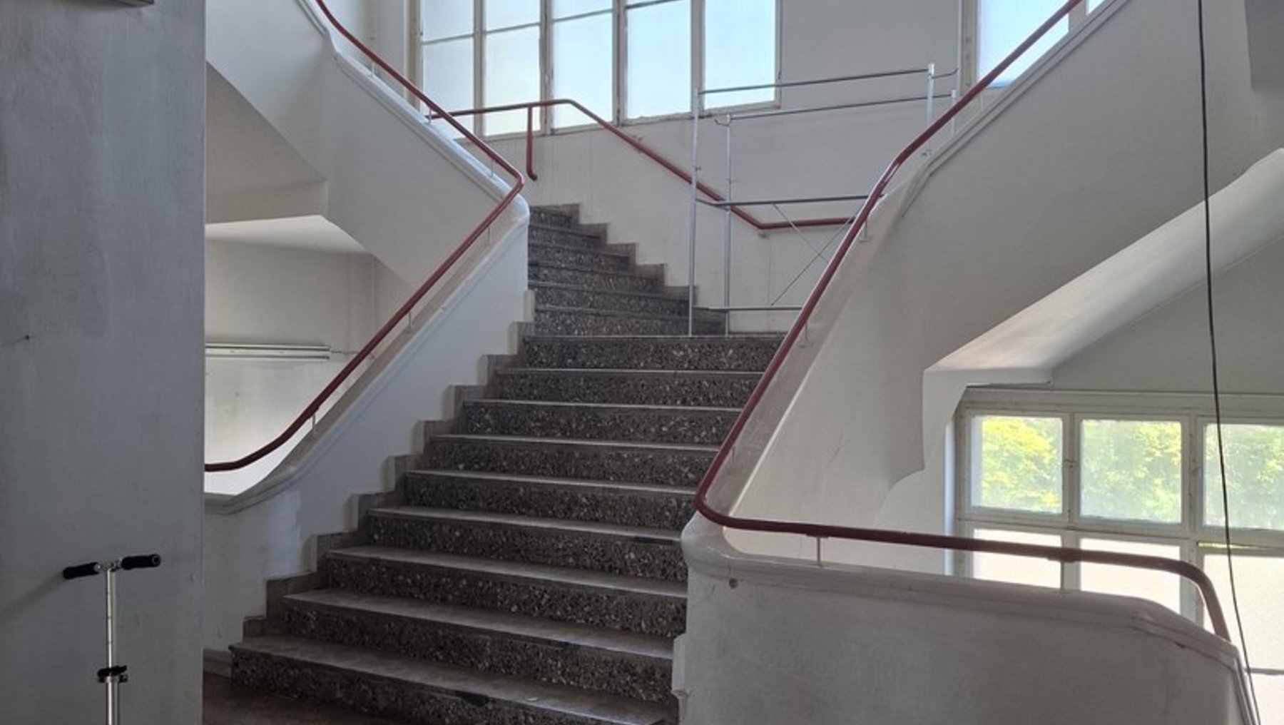 Zwei breite Treppenläufe mit Metallgeländern in einem hellen, modernen Innenraum, die zu einem oberen Stockwerk führen. Große Fenster lassen Tageslicht ein.