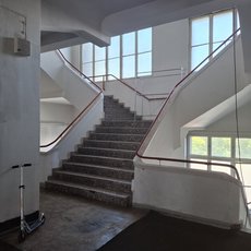 Zwei breite Treppenläufe mit Metallgeländern in einem hellen, modernen Innenraum, die zu einem oberen Stockwerk führen. Große Fenster lassen Tageslicht ein.