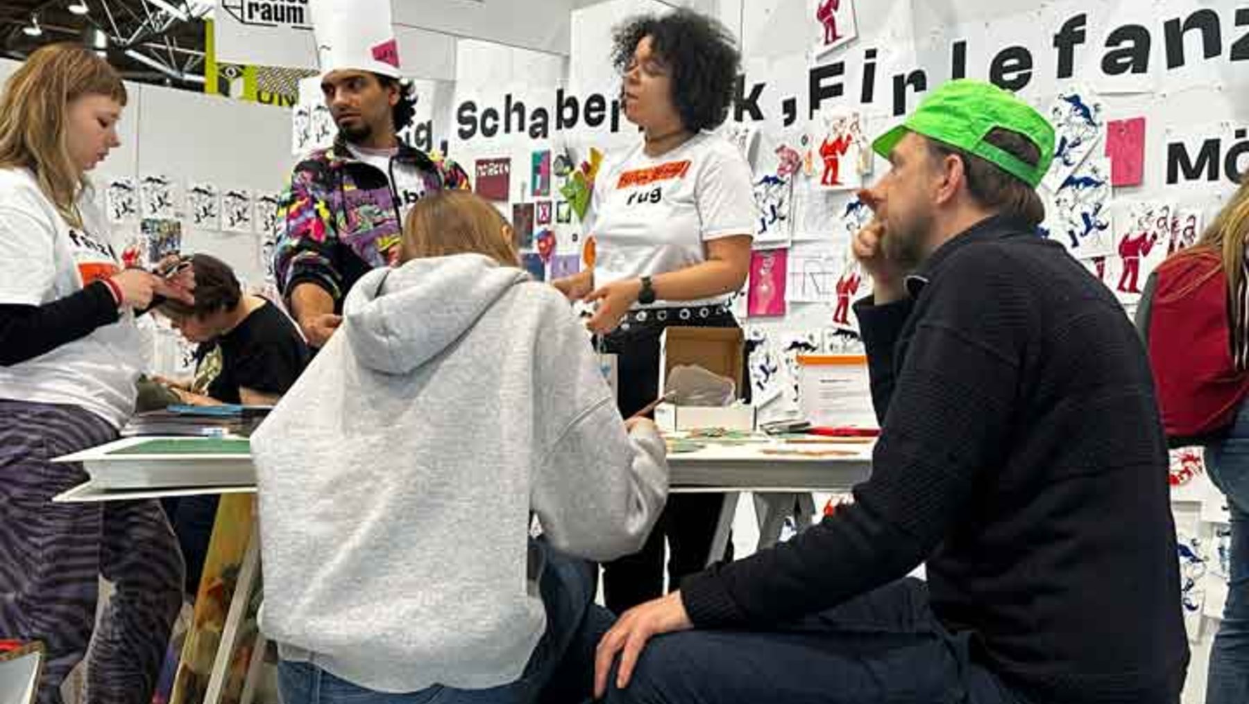 Eine Gruppe von fünf Personen unterhält sich an einem Tisch in einem kreativen Raum mit Plakaten und Postern im Hintergrund, die mit dem Titel „Schabernack, Fiplefanz“ und Illustrationen bedruckt sind.
