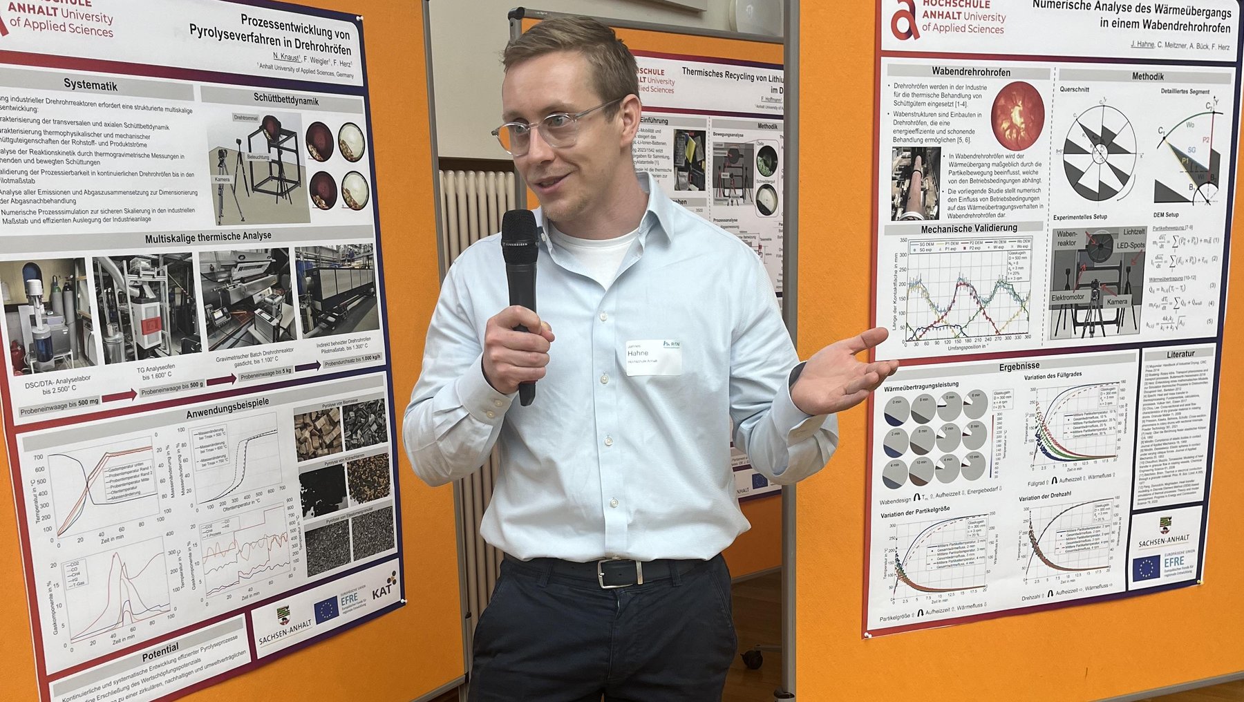 Mann hält vor zwei wissenschaftlichen Postern mit Diagrammen, Grafiken und Texten auf orangefarbenem Hintergrund eine Präsentation. Er steht mit Mikrofon und zeigt auf die Inhalte, die technische oder medizinische Themen behandeln könnten.