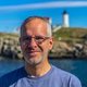 Mann mit Brille lächelt leicht, trägt ein blaues T-Shirt, vor ihm im Hintergrund ein Leuchtturm auf felsiger Küste mit blauem Himmel und Wasser.